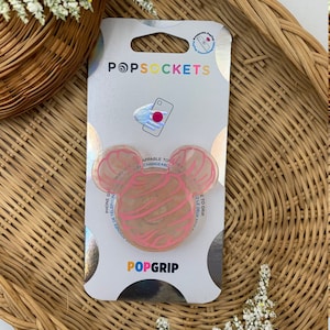 Puede incluir: Un PopGrip para teléfono rosa y blanco con un diseño de Mickey Mouse. La empuñadura está empaquetada en una tarjeta plateada y blanca con el logotipo de PopSockets.