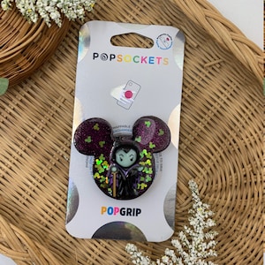 May include: A purple and green glitter phone grip with a cartoon image of Maleficent from Disney's Sleeping Beauty. The grip is a PopGrip brand and is designed to be swappable and interchangeable. The packaging says "PopSockets" and "PopGrip".