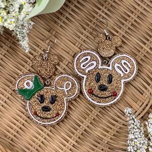 May include: A pair of gold glitter Mickey Mouse earrings with green bows. The earrings are on a woven brown background.
