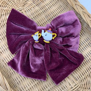 Könnte beinhalten: Eine große, tiefviolette Samtschleife mit einer Cartoon-Teekannen- und Tassenverzierung. Die Teekanne und die Tasse sind weiß mit gelben und lila Akzenten. Die Schleife ist etwa 15 cm breit.