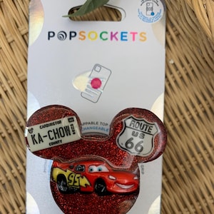 Puede incluir: Agarre para teléfono de glitter rojo con un coche de carreras rojo de dibujos animados, un cartel de la Ruta 66 y una matrícula que dice "Ka-Chow!"  Condado de Carburador.  El agarre para teléfono es una marca PopGrip.