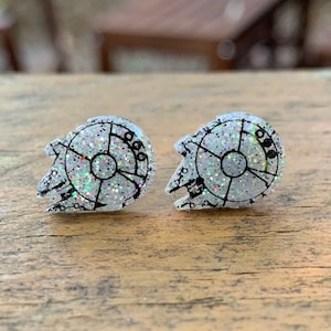 Pode incluir: Um par de brincos de pressão com glitter prateado, com um contorno branco do Millennium Falcon, a nave espacial de Star Wars.