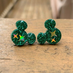 May include: A pair of green glitter earrings shaped like Mickey Mouse ears with gold glitter stars.