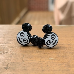 May include: A pair of black and white Mickey Mouse ear-shaped earrings with a glitter finish. The ears have a skull and crossbones design.
