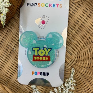 May include: A blue and green PopSocket phone grip with a glitter finish. The grip features a cloud design with the words "Toy Story" in red and yellow letters. The PopSocket is packaged on a white card with the PopSockets logo and the words "Appliable to Changeable" and "PopGrip".