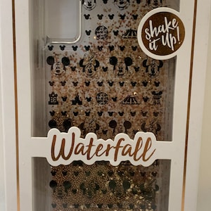 May include: A clear phone case with a gold glitter waterfall design featuring Mickey Mouse and Minnie Mouse silhouettes, along with Disney park icons. The case has a "Shake it Up!" label and says "Waterfall" on the bottom. It also states "10 Foot Drop Protection" and "for | pour".