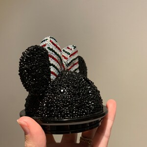 May include: Black rhinestone Minnie Mouse head with a red, white and green striped bow. The head is attached to a black base.