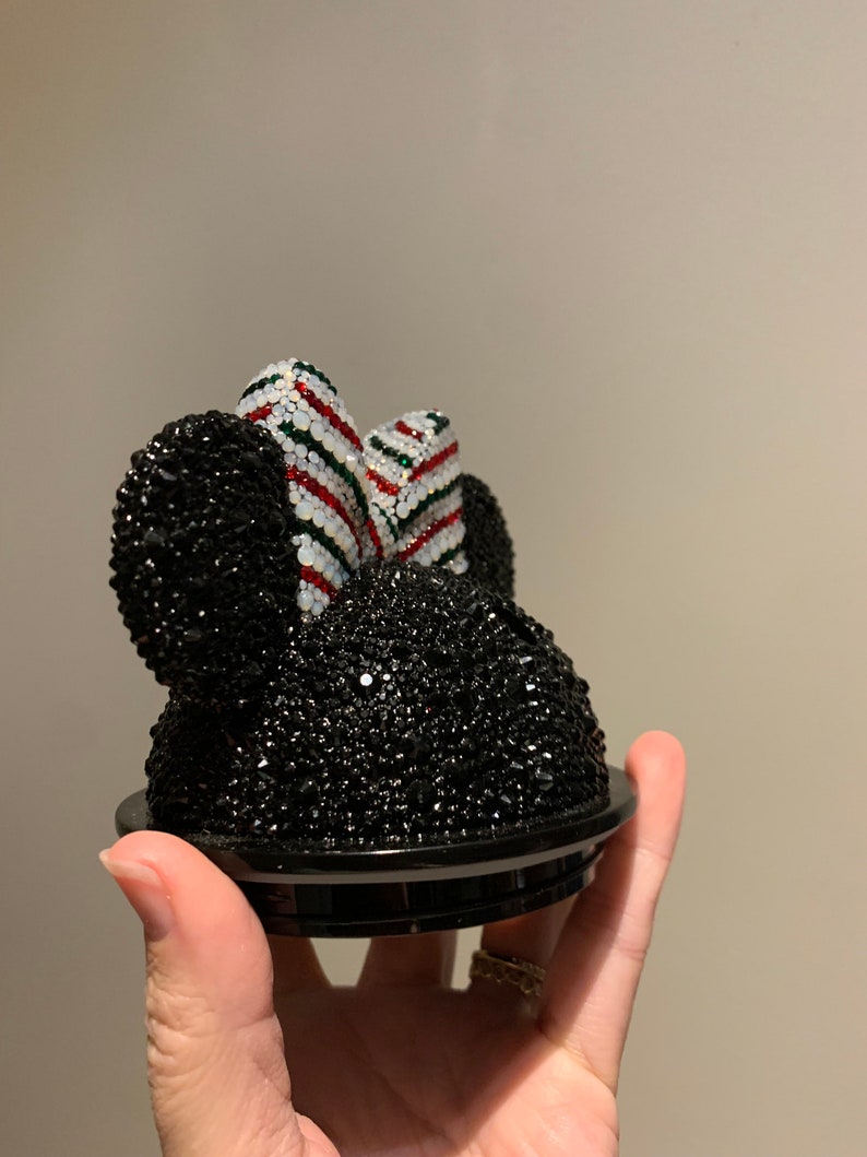 May include: Black rhinestone Minnie Mouse head with a red, white and green striped bow. The head is attached to a black base.