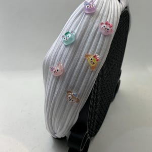 May include: White ribbed headband adorned with colorful cartoon character embellishments. The headband features a knotted design and is displayed on a black stand. The characters include a bear, a bunny, and a cat.