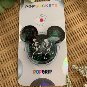 Puede incluir: Un agarre para teléfono de color negro y verde con purpurina y un diseño de esqueleto blanco. El agarre tiene forma de orejas de Mickey Mouse. El embalaje dice "PopSockets" y "PopGrip".