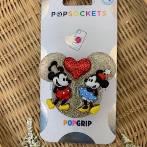May include: A PopSockets PopGrip featuring Mickey Mouse and Minnie Mouse in a glitter heart design. The PopGrip is white with a red glitter heart in the center. Mickey Mouse is wearing red shorts and yellow shoes. Minnie Mouse is wearing a blue and white polka dot dress and yellow shoes. The PopGrip is packaged on a white card with the PopSockets logo.