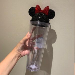 May include: A clear plastic tumbler with a black and red rhinestone Minnie Mouse lid. The lid features a black bow with red rhinestones.