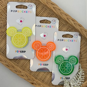 May include: Three phone grips with a Mickey Mouse shaped design. The grips are yellow, orange, and green and feature a lemon slice design. The packaging says "PopSockets" and "PopGrip".