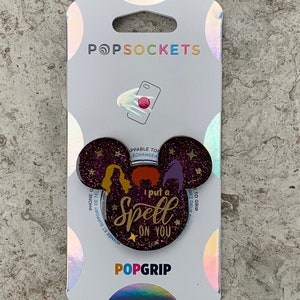 Puede incluir: Una agarradera de teléfono de purpurina morada con una silueta de Mickey Mouse. La agarradera presenta un gráfico de tres brujas con el texto "I put a spell on you".