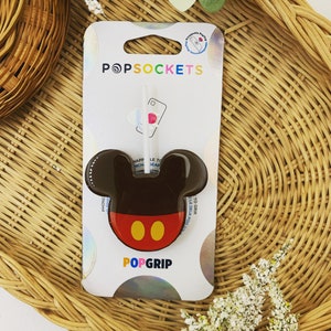 May include: A brown and red PopGrip phone stand with a Mickey Mouse design. The PopGrip is on a white card with the words "PopSockets" and "PopGrip" printed on it.