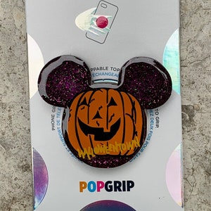May include: A purple and black glitter phone grip with a pumpkin shaped design. The pumpkin is orange with a black jack-o-lantern face and the text "HALOWEENTOWN" in yellow letters. The phone grip is labeled "POPGRIP".
