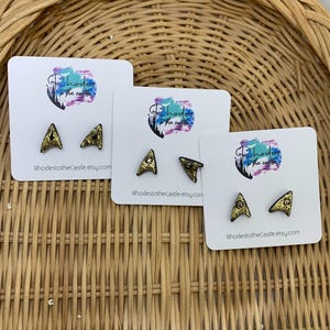 May include: Three pairs of gold glitter Star Trek inspired earrings on white cards with the text "Rhodes to the Castle" and the website address "RhodestotheCastle.etsy.com".