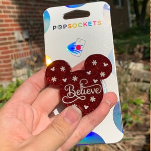 May include: Red glitter phone grip with a Mickey Mouse silhouette and the word "Believe" in white. The grip is on a white card with the word "PopSockets" printed on it.