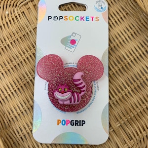 May include: Pink glitter PopSockets PopGrip featuring a smiling Cheshire Cat design. The PopGrip is in the shape of Mickey Mouse ears. The packaging includes the PopSockets and PopGrip logos.