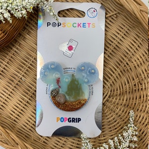 May include: A blue and green PopSockets PopGrip featuring a mermaid, seashells, and glitter. The text "PopGrip" and "Appliable to Changeable" are printed on the packaging.