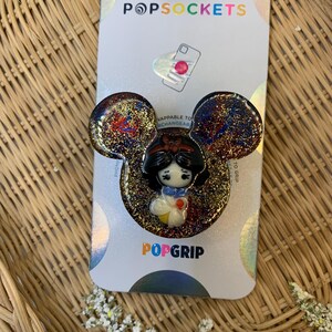 May include: A PopGrip phone accessory featuring a glitter-filled design with a Snow White character inside. The PopGrip is compatible with PopSockets and is designed to be interchangeable.