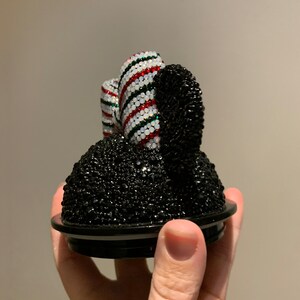 May include: A black and white jeweled Mickey Mouse ear-shaped lid for a container. The ear is covered in black rhinestones and the bow is covered in white, red, and green rhinestones.