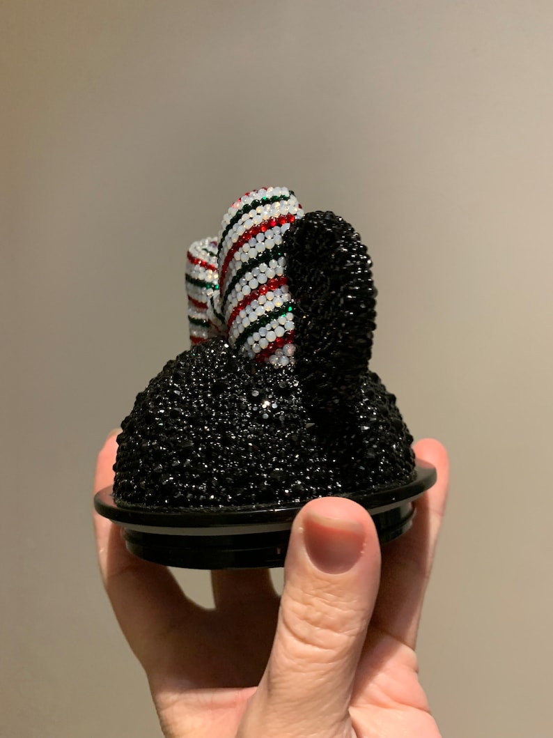 May include: A black and white jeweled Mickey Mouse ear-shaped lid for a container. The ear is covered in black rhinestones and the bow is covered in white, red, and green rhinestones.