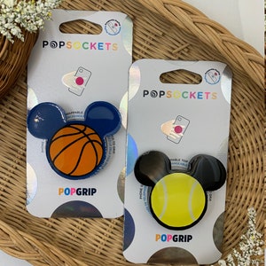 May include: Two PopSockets phone grips with a Mickey Mouse design. The first grip features a blue ear with a basketball in the center. The second grip features a black ear with a tennis ball in the center. Both grips have the text "PopGrip" printed on the bottom.
