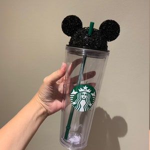 May include: A clear plastic tumbler with a black glitter Mickey Mouse lid and a green Starbucks logo. The tumbler has a green straw.