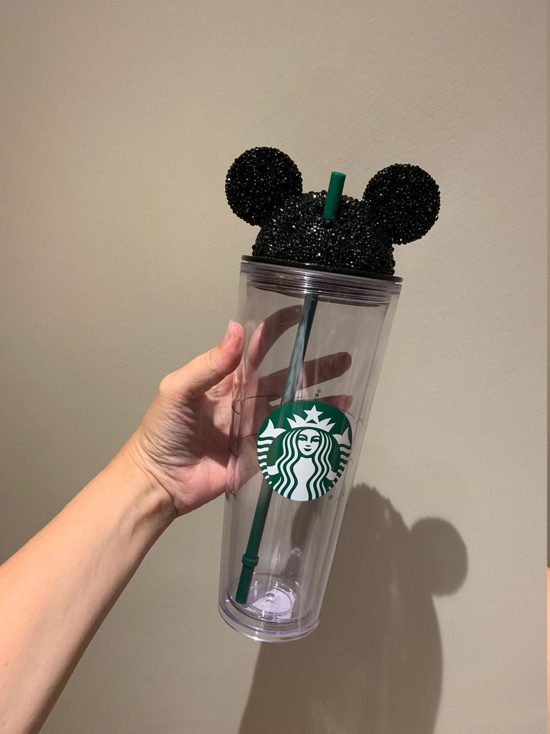 May include: A clear plastic tumbler with a black glitter Mickey Mouse lid and a green Starbucks logo. The tumbler has a green straw.