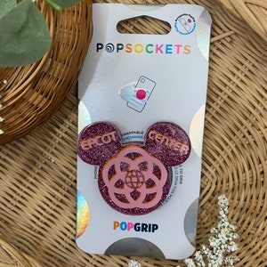 Puede incluir: Un agarre para teléfono PopGrip rosa y naranja con un acabado brillante. El agarre presenta un diseño de cabeza de Mickey Mouse con las palabras "Epcot" y "Center" en las orejas. El centro del agarre presenta un diseño rosa y naranja con un globo en el centro.