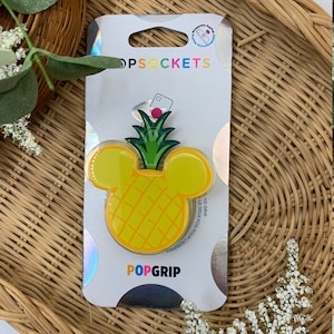 May include: A yellow pineapple-shaped PopGrip phone stand with a green top and Mickey Mouse ears. The PopGrip is packaged on a white card with the words "PopSockets" and "PopGrip" printed on it.