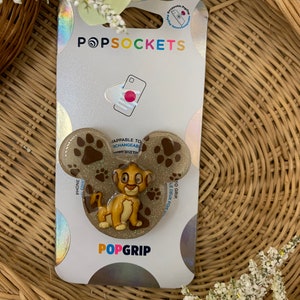 May include: A gold glitter phone grip with a paw print design and a cartoon image of Simba from The Lion King. The grip is labeled "PopGrip" and is compatible with PopSockets.