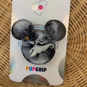 May include: A black and white PopGrip phone grip with a glittery finish. The grip features a white silhouette of the character Zero from the movie The Nightmare Before Christmas. The text "POPGRIP" is printed in blue, red, and yellow on the packaging.