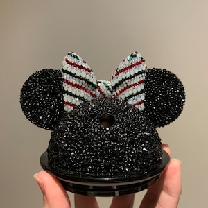 May include: Black and white Minnie Mouse ears with a red and green striped bow. The ears are covered in rhinestones.