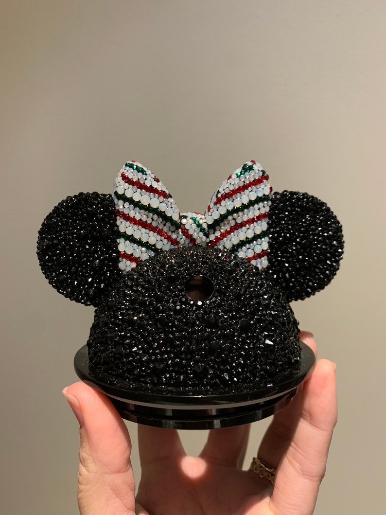 May include: Black and white Minnie Mouse ears with a red and green striped bow. The ears are covered in rhinestones.