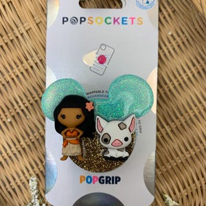 May include: A turquoise and gold glitter PopGrip phone accessory featuring Moana and Pua from the Disney movie Moana. The PopGrip is a reusable and swappable phone grip.