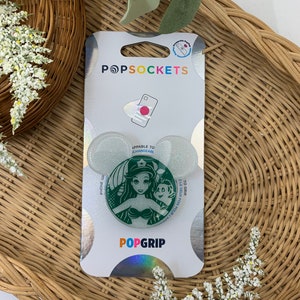 May include: A green and white PopGrip phone grip featuring a glitter background and a graphic of Ariel from The Little Mermaid with Flounder. The text "PopGrip" is printed in white on the grip. The packaging is white with the text "PopSockets" printed in black.