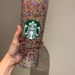 May include: A clear plastic tumbler with a green Starbucks logo and a colorful glitter design. The glitter includes stars, flowers, and small black and white shapes.