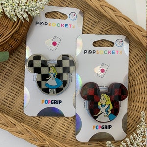 Può includere: Due impugnature per telefono PopSockets con Alice nel Paese delle Meraviglie. L'impugnatura a sinistra è a scacchi bianco e nero con uno sfondo di glitter argentato. L'impugnatura a destra è a scacchi rosso e nero con uno sfondo di glitter rosso. Entrambe le impugnature hanno la forma di una testa di Topolino e il personaggio di Alice nel Paese delle Meraviglie.