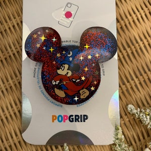 Puede incluir: Un agarre para teléfono de color rojo y azul con purpurina con un personaje de Mickey Mouse vestido de mago. El agarre tiene un patrón de estrellas y el texto "PopGrip" está impreso en la parte inferior.