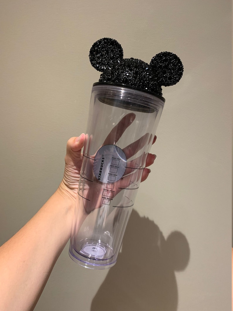 May include: Clear plastic reusable Starbucks tumbler with a black Mickey Mouse ears lid covered in black rhinestones. The tumbler has measurement markings for sugar, cream, shots, milk, and total ounces.
