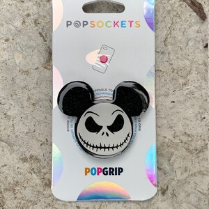 May include: Black and white PopSocket phone grip featuring a Jack Skellington character from the Nightmare Before Christmas movie. The PopSocket is on a white background with the words "PopSockets" and "PopGrip" printed on the packaging.