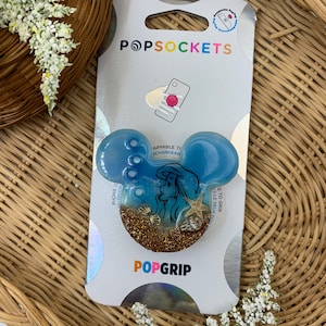 May include: A blue and gold glitter phone grip with a mermaid illustration. The grip is compatible with PopSockets and is labeled "PopGrip".