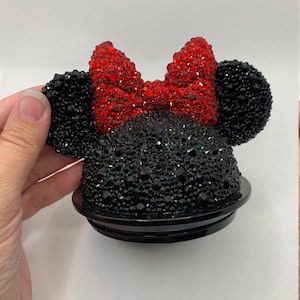 May include: Black and red rhinestone Minnie Mouse-themed bottle stopper. The stopper features a black base with a red bow and black ears, all covered in rhinestones.