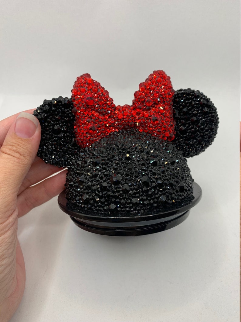 May include: Black and red rhinestone Minnie Mouse-themed bottle stopper. The stopper features a black base with a red bow and black ears, all covered in rhinestones.