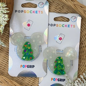 May include: Two white PopSockets phone grips with a glittery finish. The grips feature a green Christmas tree design with colorful ornaments and Mickey Mouse heads. The text "PopGrip" is printed on the bottom of each grip.
