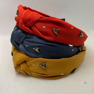 Puede incluir: Tres diademas anudadas en rojo, azul y amarillo con adornos de estrellas y triángulos dorados.