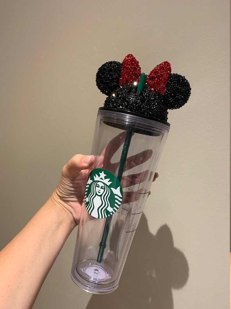 May include: A clear plastic tumbler with a black and red rhinestone Minnie Mouse lid. The tumbler has a green Starbucks logo on the side.