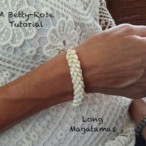 Op de afbeelding: Een witte kralen armband met een zilveren sluiting. De armband is gemaakt van lange, smalle kralen die in een patroon zijn gerangschikt. De armband wordt om de pols gedragen.